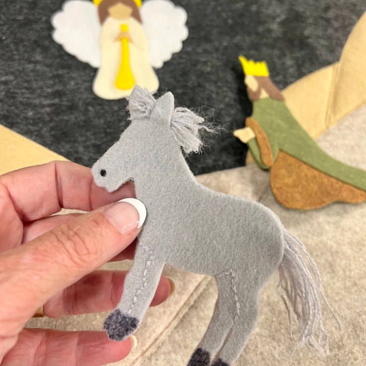 A hand holds a small gray felt donkey. In the background, felt figures of an angel and a person wearing a crown are visible, suggesting a nativity or Christmas scene.