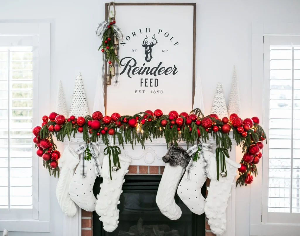 A white fireplace mantle showcases festive christmas mantel decorating with red ornaments, green garland, white stockings, and cone trees. Above, a "North Pole Reindeer Feed" sign is adorned with a charming bunch of greenery.