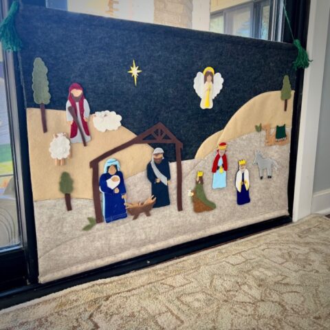 A felt nativity scene with figures of Mary, Joseph, baby Jesus, three wise men, an angel, a shepherd, a sheep, and a donkey, set against a dark background with a yellow star.