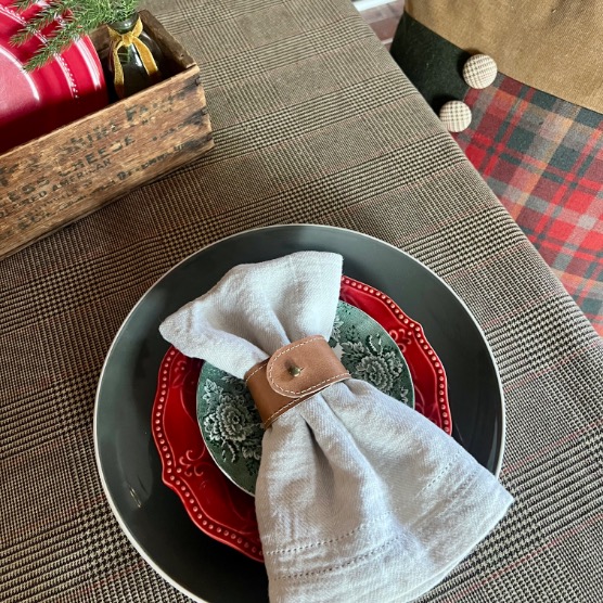 A festive table setting inspired by Ralph Lauren Christmas ideas features a gray plaid tablecloth, a green and red plate, and a napkin in a brown leather ring. In the background, part of a wooden box and someone wearing plaid can be seen.