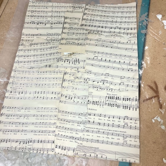 A sheet of music made from multiple smaller pieces of sheet music taped together, forming a patchwork pattern. The sheet is on a work surface with paint splatters and a blue tool to the right.