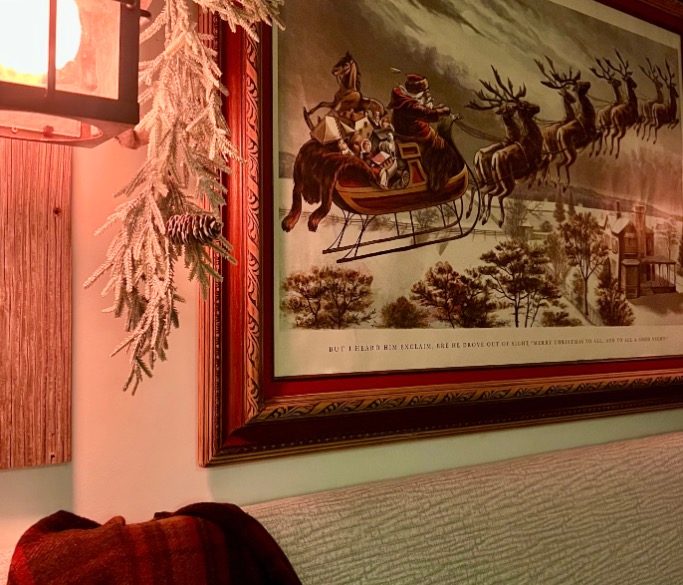 A framed vintage-style print of Santa Claus in a sleigh pulled by reindeer hangs on a wall. Pine branches and a glowing lantern add a cozy, festive touch to the scene. A blanket is draped over the back of a couch below.