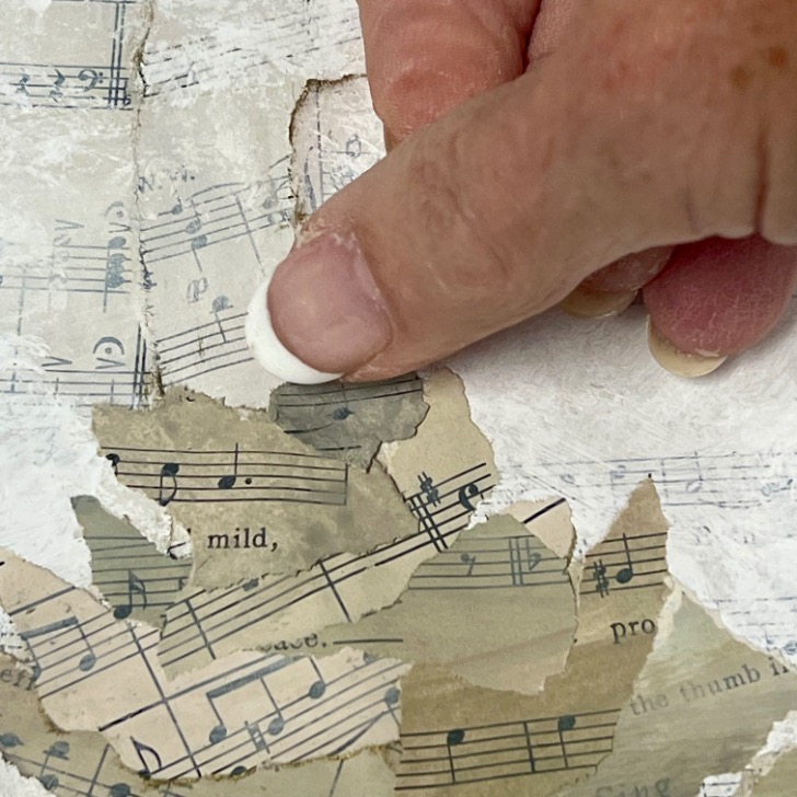 A hand with short, clean fingernails presses down on overlapping torn pieces of sheet music paper, arranged on a textured white surface.