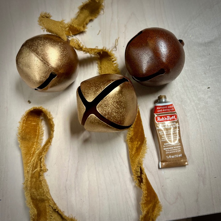 Three large jingle bells, two gold and one bronze, rest on a light surface with frayed golden ribbon and a small tube of Rub’n Buff metallic finish nearby—perfect for elegant Ralph Lauren Christmas ideas.