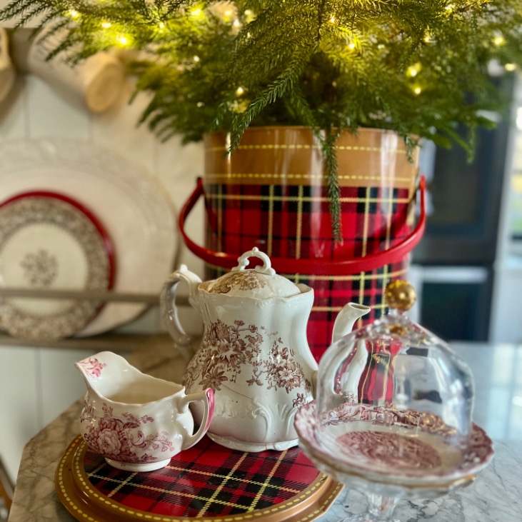 A festive holiday table features a red plaid tin with evergreen branches, echoing Ralph Lauren Christmas ideas, along with a decorative teapot, sugar bowl, and creamer with red patterns. Warm lights add a cozy touch to this inviting scene.