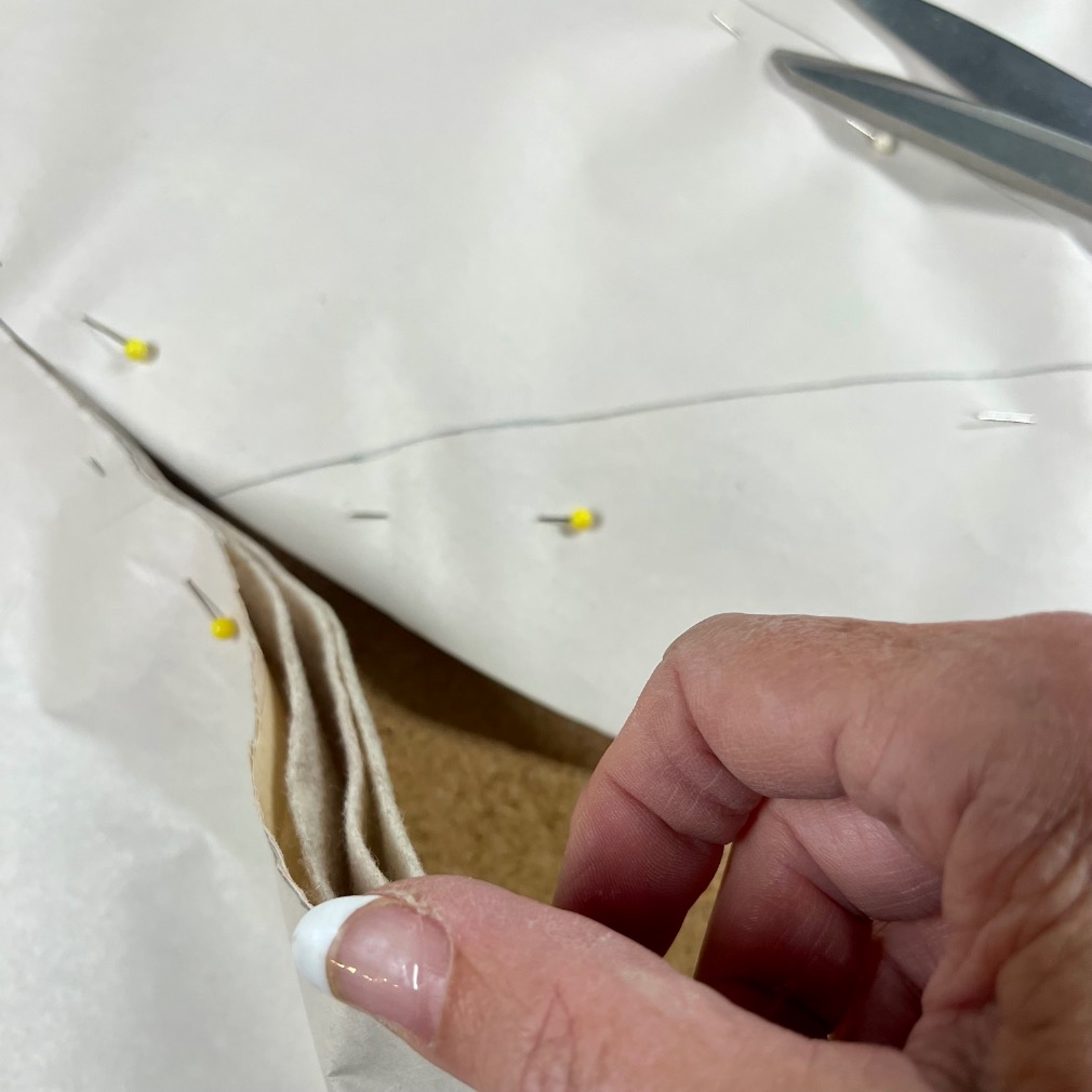 A hand with a French manicure holds folded layers of beige fabric pinned together with yellow-headed pins. Scissors and more pins are visible on a white surface in the background.