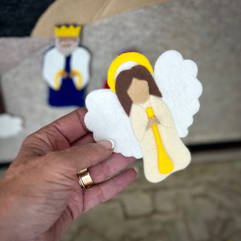 A hand holding a felt angel with white wings, a yellow halo, and a yellow trumpet; a felt king figure is blurred in the background.