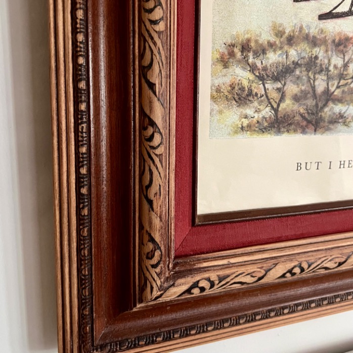 Close-up of the corner of a framed artwork with an ornate, carved wooden frame and a red matte border; part of a landscape painting and the printed words "BUT I HE" are visible, evoking classic Ralph Lauren Christmas ideas.