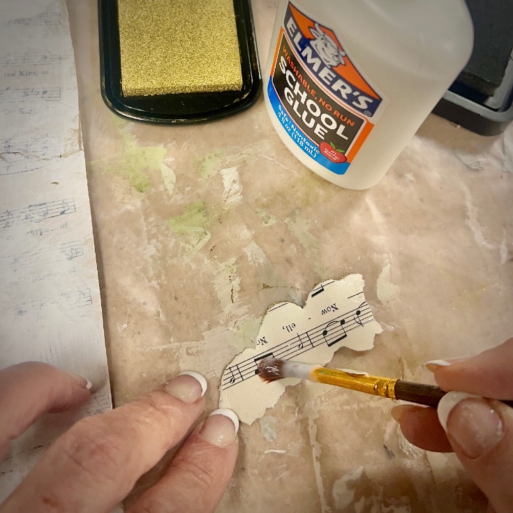 A person uses a small paintbrush to apply glue to a torn piece of sheet music on a table, with a bottle of Elmer’s School Glue and a sponge in the background.