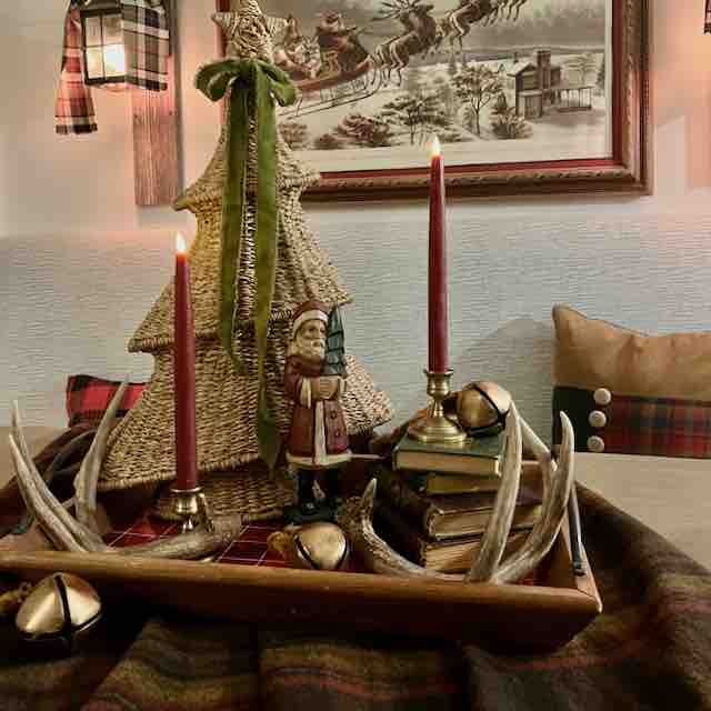 A cozy holiday centerpiece features red candles, a woven tree, a Santa figurine, gold bells, stacked books, and antlers on a tray—plaid accents and a Santa sleigh painting evoke timeless Ralph Lauren Christmas ideas.