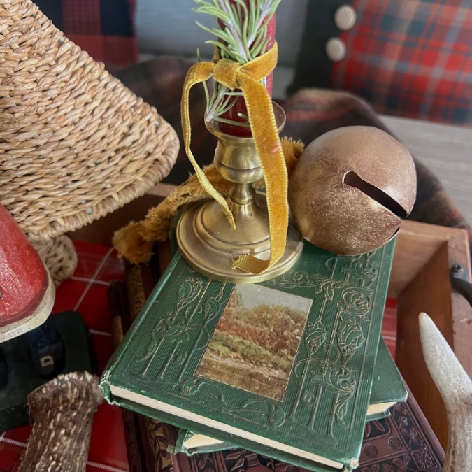 A green, embossed book with a landscape illustration sits on a tray, topped with a brass candlestick wrapped in yellow ribbon, an artificial plant, and a large bronze jingle bell—evoking cozy Ralph Lauren Christmas ideas among the rustic decor.