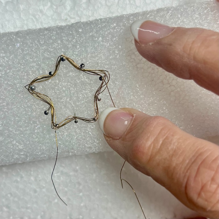A hand holds a delicate wire shaped like a star, decorated with tiny beads, against a piece of white foam. The wire is partially threaded with thin strands.