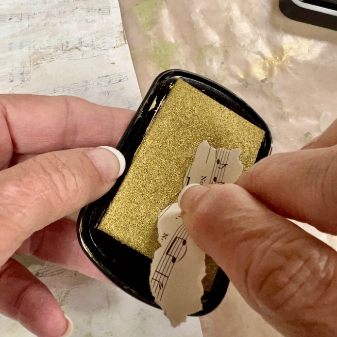 A person rubs a torn piece of sheet music onto a gold ink pad, preparing to apply ink for a craft project.