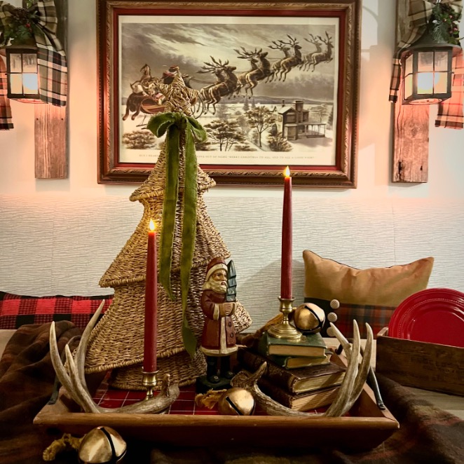 A cozy holiday display inspired by Ralph Lauren Christmas ideas features a woven Christmas tree, red candles, a Santa figurine, books, gold bells, and antlers on a tray. Plaid accents and lantern light complete the inviting scene.