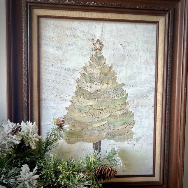 A framed artwork of a Christmas tree made from layered sheet music, topped with a star. Pine branches with white-tipped needles and pinecones are arranged in front of the frame.