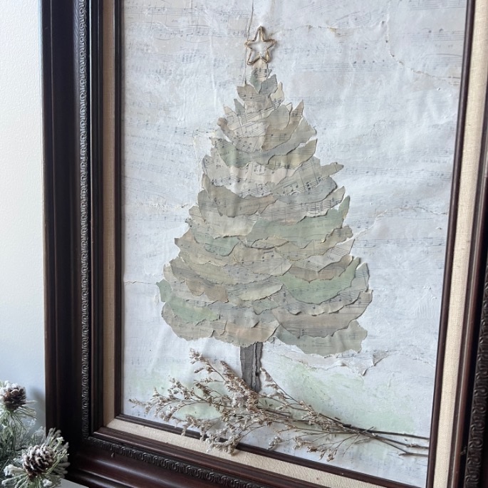 Framed artwork of a Christmas tree made from layered, textured paper with a star on top, set against a light background. Pinecones and evergreen branches decorate the lower left corner of the frame.