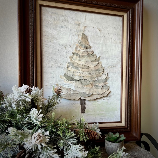 Framed art of a Christmas tree made from layered, torn paper hangs on a wall. In front, a pine branch with frosted needles and pinecones, and a small potted succulent sit on a table.