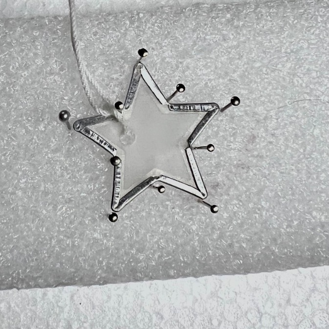 A silver star-shaped ornament with a white center and metallic points is hanging by a white string against a textured white background.
