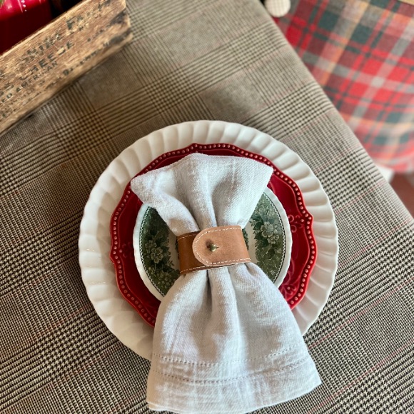 A white napkin with a brown leather ring rests on green and white plates stacked on a red and white scalloped plate, set on a plaid tablecloth—perfect inspiration for Ralph Lauren Christmas ideas. A plaid chair and a wooden box are partially visible nearby.