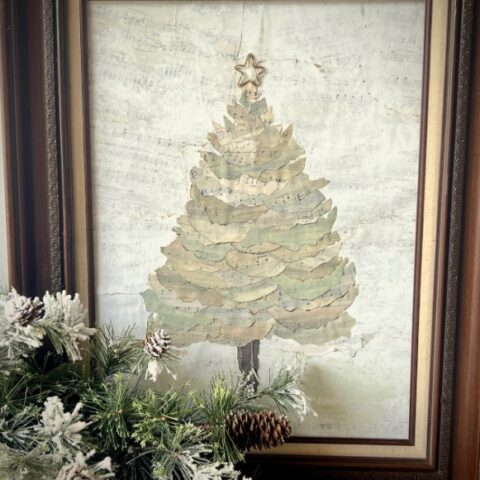 Framed artwork of a Christmas tree made from layered torn sheet music, topped with a star. In front, a snowy pine branch with pinecones partially covers the bottom left corner of the frame.