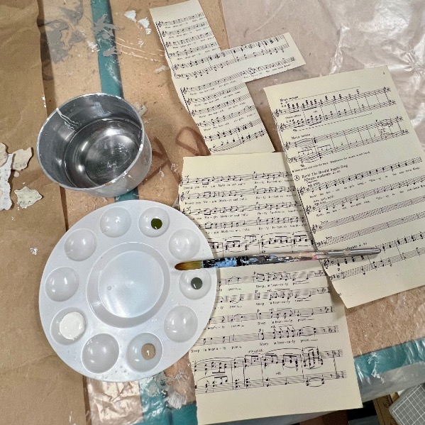 A paint palette, paintbrush, bowl of water, and several sheets of printed sheet music are spread out on a workspace covered with brown paper and plastic. Some sheets have been cut into shapes.