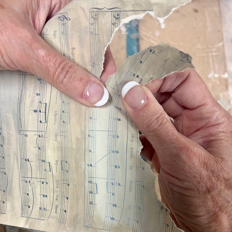 Two hands with manicured nails are tearing a sheet of printed music over a beige surface. The sheet music shows notes and lyrics.