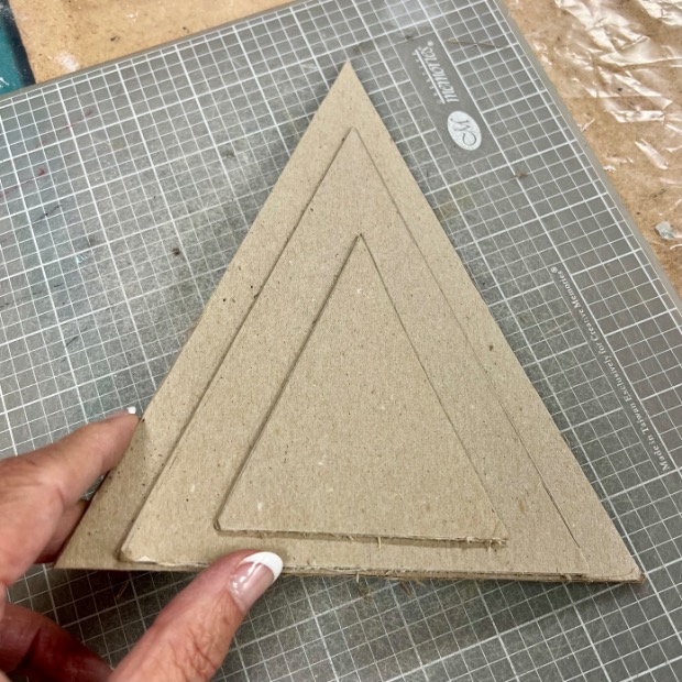 A hand holds a triangular piece of cardboard with two smaller, concentric triangles outlined inside it. The piece rests on a cutting mat with a grid and measurement markings.