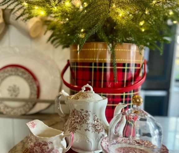 A festive holiday scene with a plaid bucket of evergreen branches and string lights, next to vintage red and white china, including a teapot, creamer, and a plate under a glass cloche.