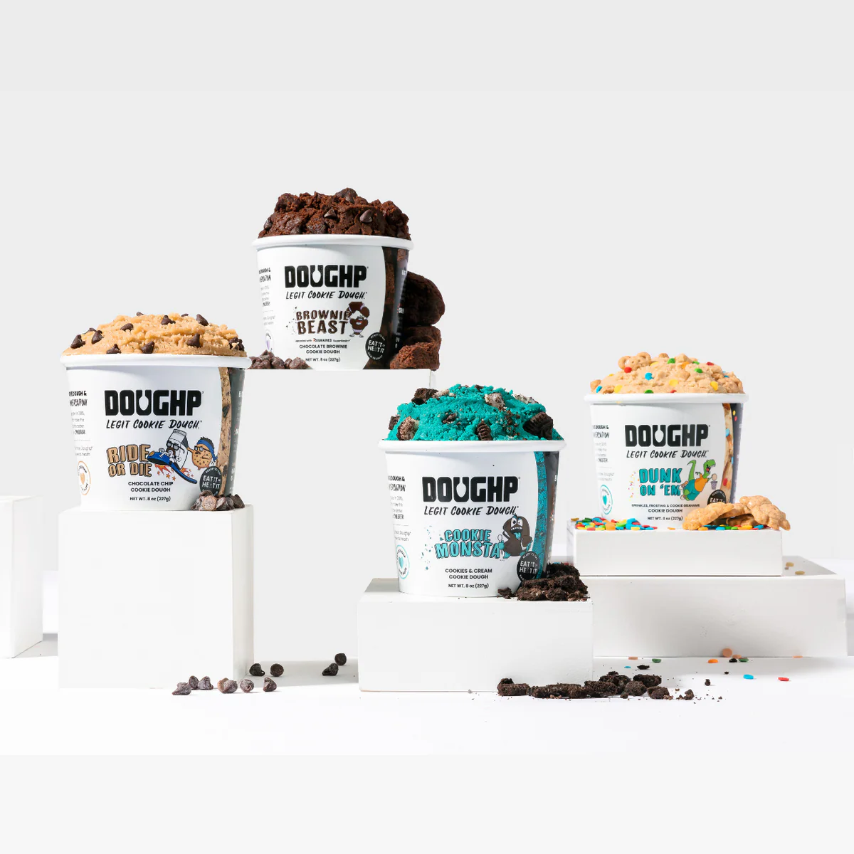 Four Doughp cookie dough tubs are displayed on white blocks, each topped with a scoop of dough in various flavors and colorful toppings, surrounded by scattered chocolate chips and cookie pieces.