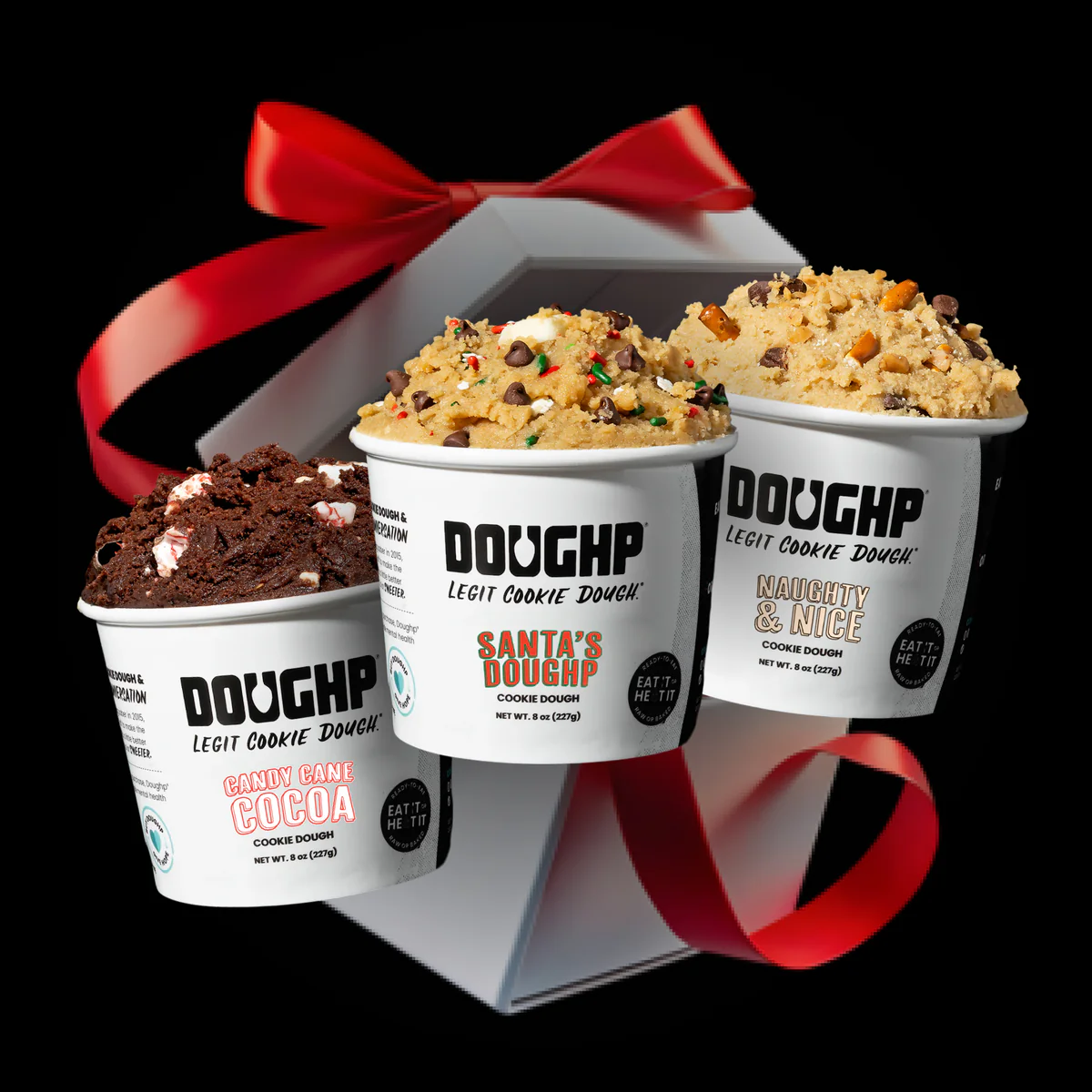 Three pints of Doughp cookie dough (Candy Cane Cocoa, Santa's Dough, Naughty & Nice) are displayed in front of a white gift box with a red ribbon bow, set against a black background.