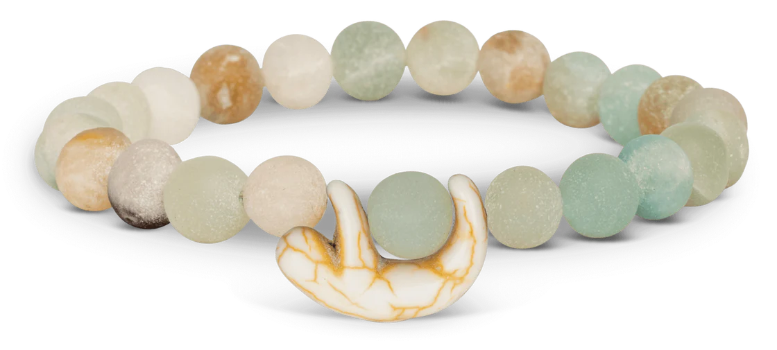 A bracelet made of round, frosted pastel beads in white, beige, and light blue tones, featuring a small, white and brown marbled sloth charm hanging from the center.