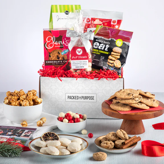 A gift box labeled "PACKED WITH PURPOSE" is filled with assorted snacks and cookies. Surrounding it are bowls and plates with popcorn, cookies, and candies, along with festive decorations like pine and ribbon.