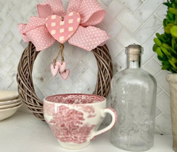 A wicker wreath with a pink bow and hanging hearts sits on a white tile backsplash. In front are a pink and white patterned teacup, a frosted glass bottle, stacked plates, and a potted green plant.