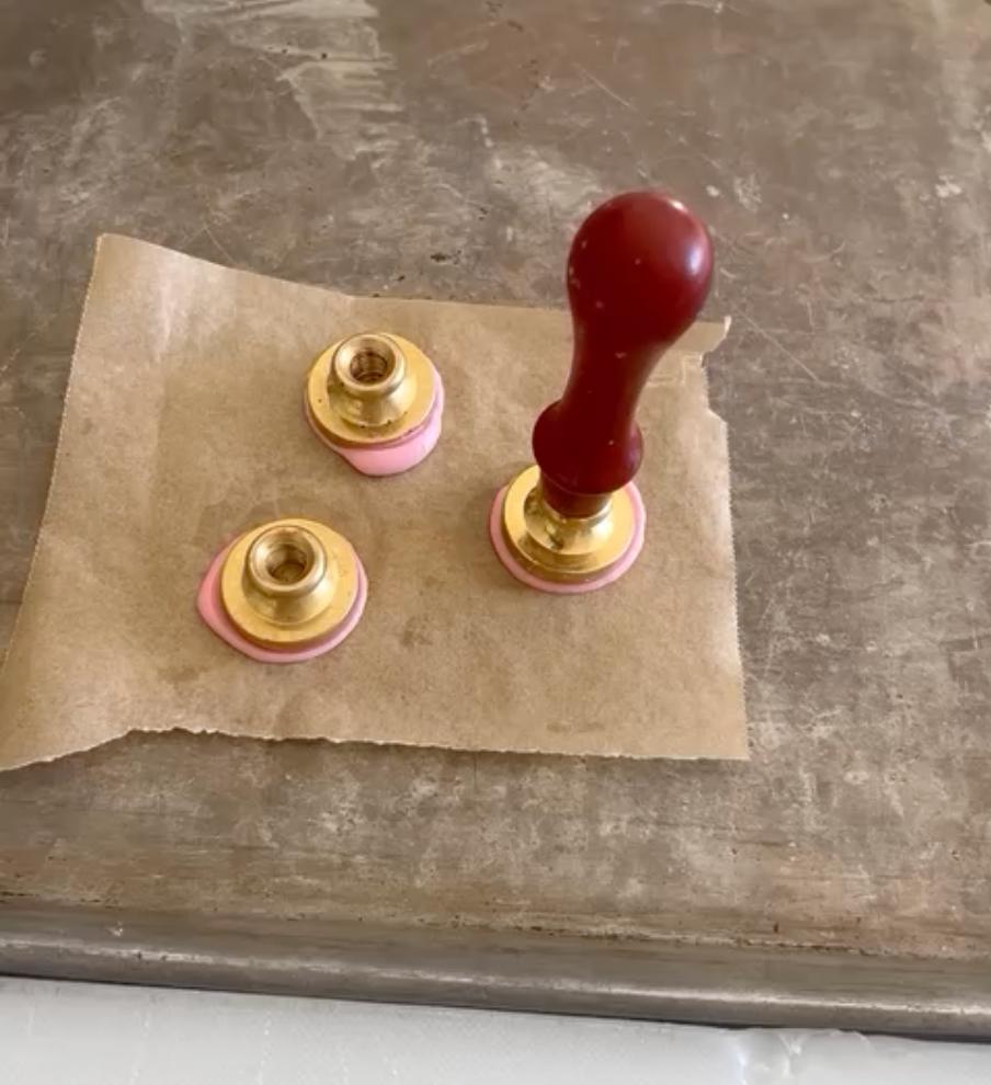 A wax seal stamp and two finished pink wax seals with brass toppers are placed on a piece of parchment paper on a metal surface.