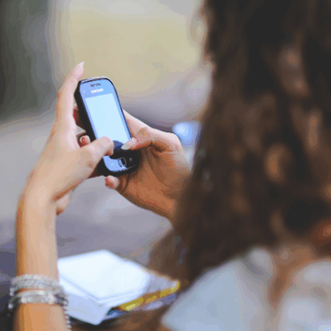 A person with long curly hair uses a smartphone outdoors, holding it in both hands. The focus is on the phone, and some papers are on the table in front of them.
