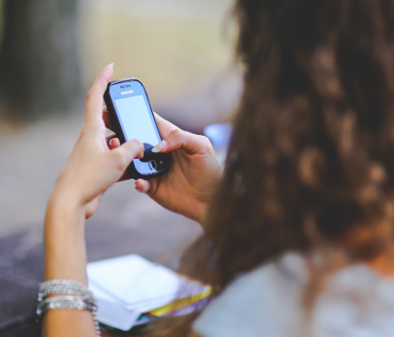 A person with long curly hair uses a smartphone outdoors, holding it in both hands. The focus is on the phone, and some papers are on the table in front of them.