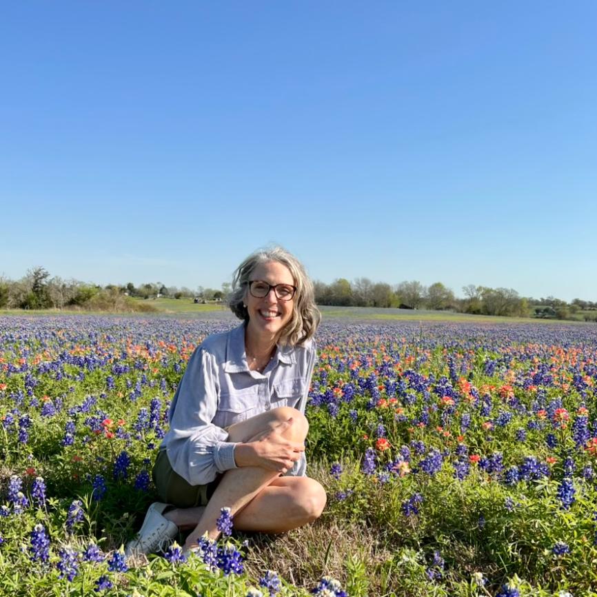 A woman with gray hair and glasses sits smiling in a field of blooming bluebonnets and wildflowers under a clear blue sky, with trees in the distance.
