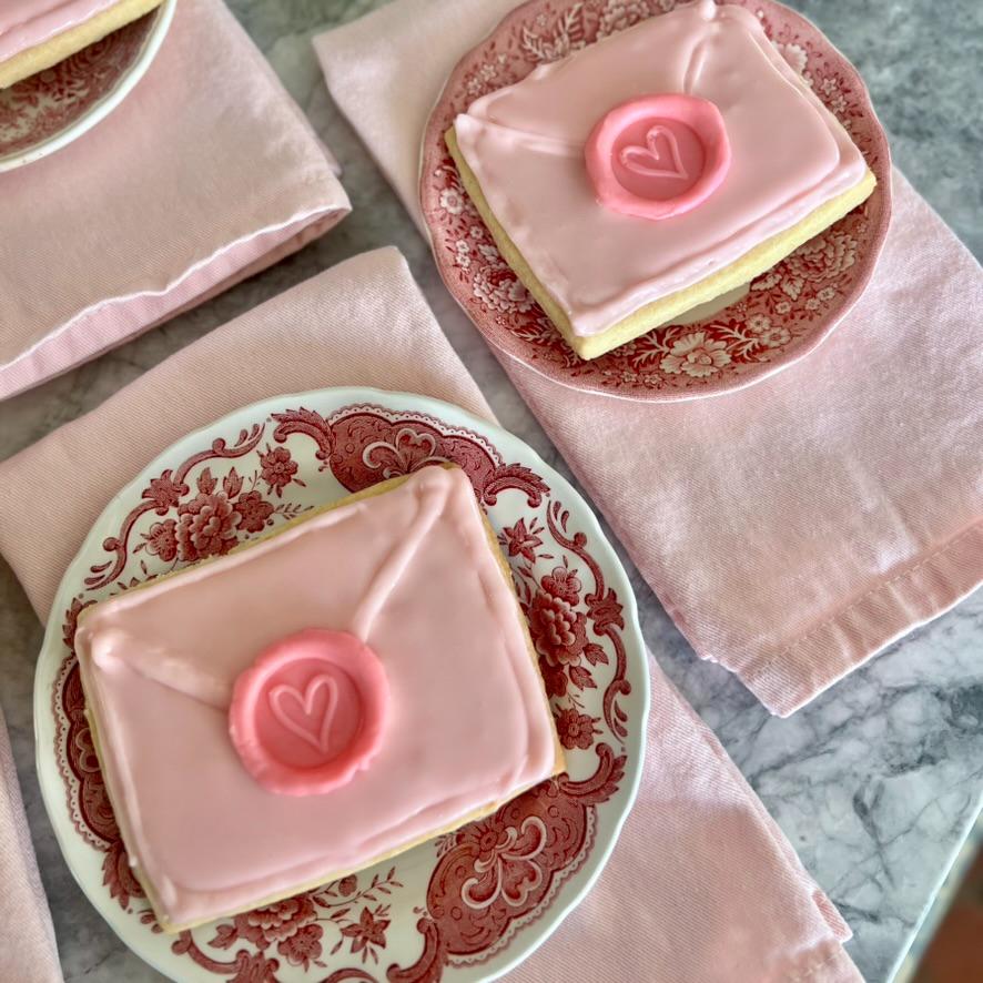 Two plates with ornate pink patterns hold cookies decorated as pink envelopes sealed with a heart. The plates rest on folded pink napkins on a marble surface, creating a soft, elegant look.