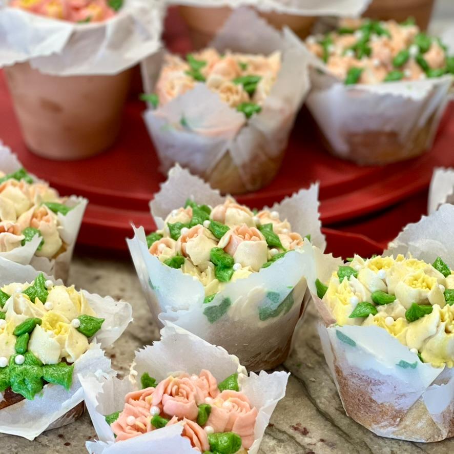 Cupcakes decorated with pastel-colored frosting shaped like flowers and leaves, wrapped in white paper, arranged on a countertop with more cupcakes on a red tray in the background.