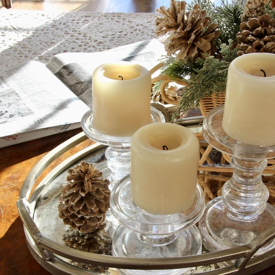 A round tray with three cream-colored pillar candles on glass holders, surrounded by pinecones and greenery. An open newspaper and a crocheted blanket are in the background on a wooden surface.