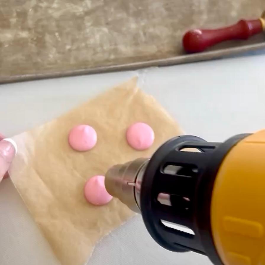 A hand holds parchment paper with three pink blobs while a heat gun is aimed at them, smoothing their surfaces. A baking tray and sealing wax tool are visible in the background.