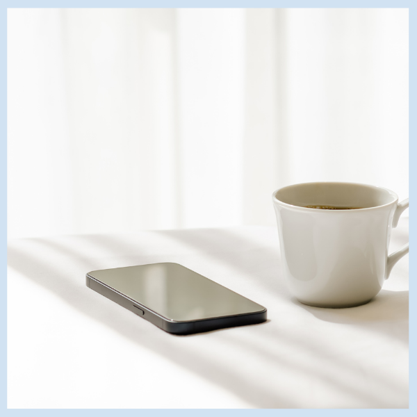 A white coffee cup and a smartphone rest on a white table, illuminated by soft natural light coming through sheer curtains in the background.