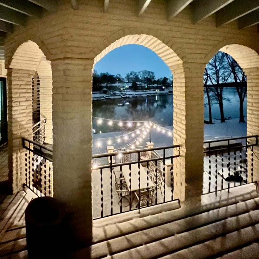 A patio overlooking a lake at dusk, seen through three brick arches. String lights hang above an outdoor dining table and chairs, with bare trees and calm water visible in the background.