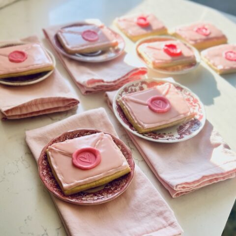 Plates with envelope-shaped cookies topped with pink icing and a heart-shaped seal sit on folded pink napkins, arranged on a light-colored table in bright sunlight.