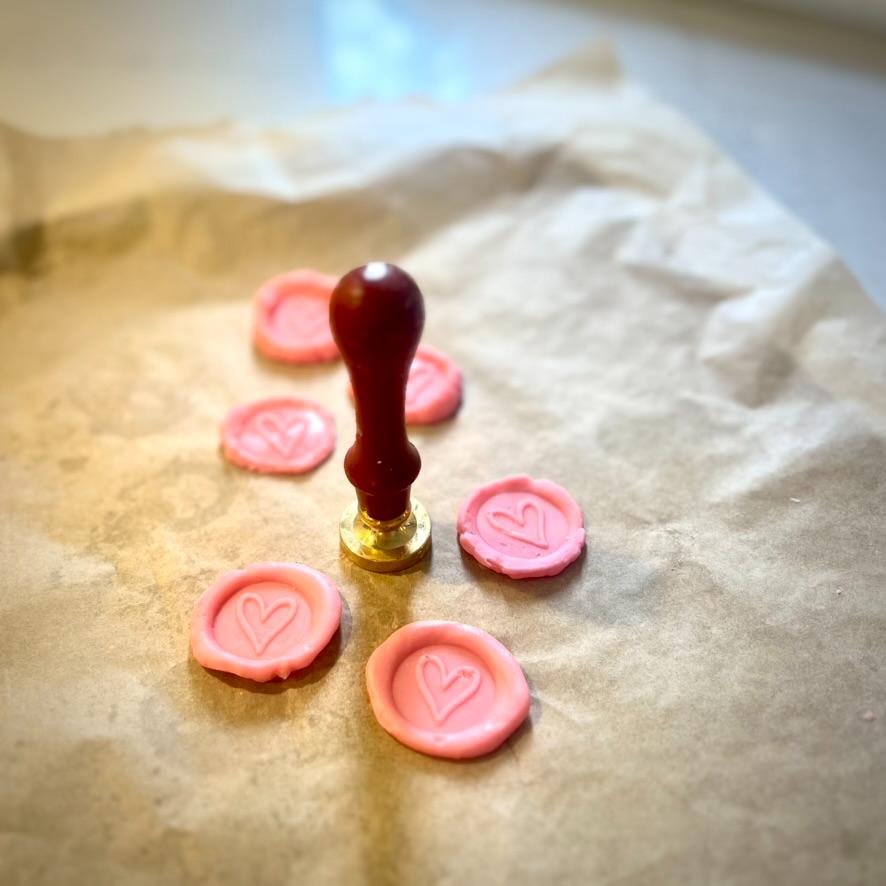 A wax seal stamp surrounded by five pink wax seals each imprinted with a heart, all placed on a sheet of crumpled parchment paper in soft, natural light.