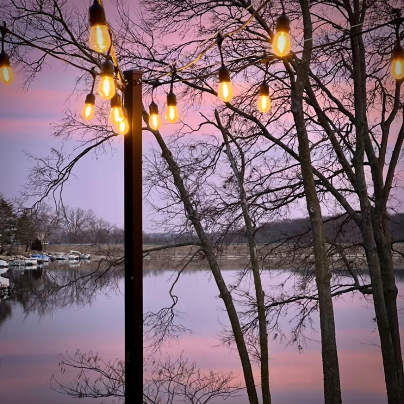 String lights hang in the foreground, glowing warmly at dusk. Leafless trees and a calm lake reflect the pink and purple hues of the sunset, creating a peaceful, scenic atmosphere.