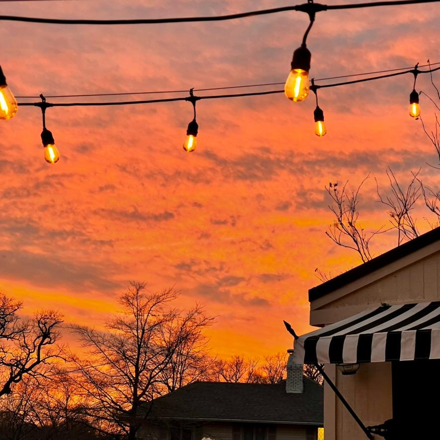 Outdoor scene at sunset with a vivid orange sky, silhouetted trees, hanging string lights, and part of a building with a black-and-white striped awning visible in the foreground.