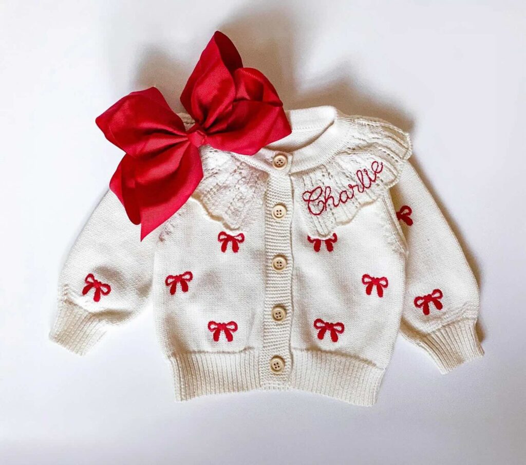 A white knitted baby cardigan with red bows embroidered all over, cream buttons down the front, “Charlie” embroidered in red on the collar, and a large red fabric bow attached to the left shoulder.