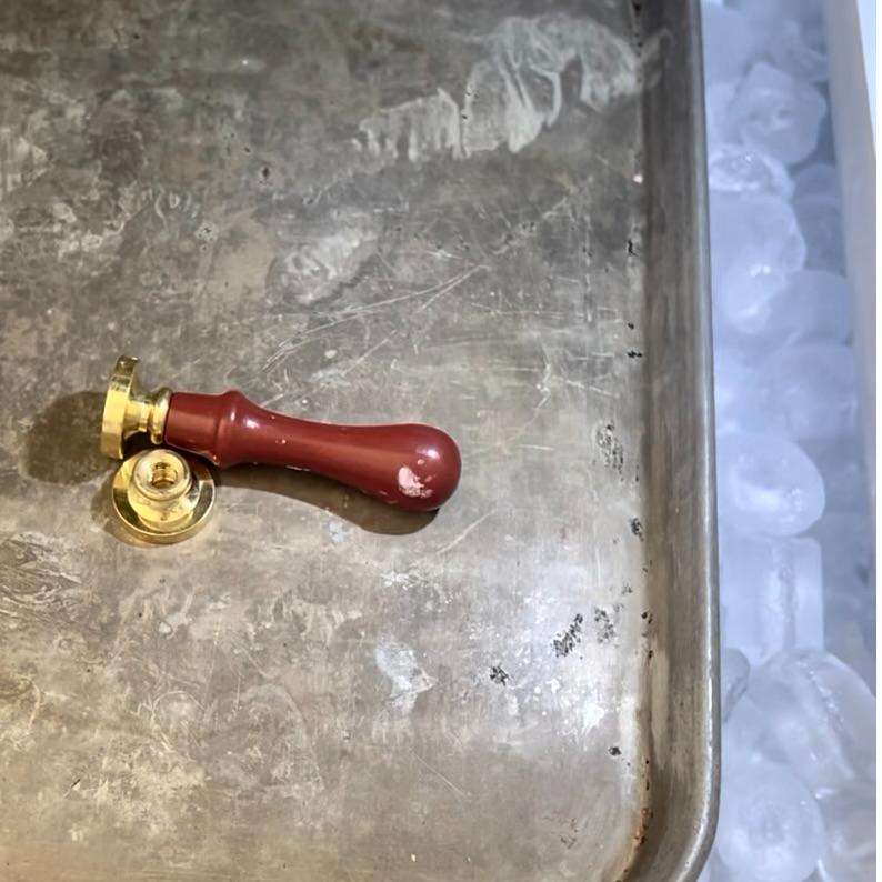 A metal tray with a red-handled brass wax seal stamp rests beside a bin filled with ice cubes. The tray has scratches and marks, showing signs of use.