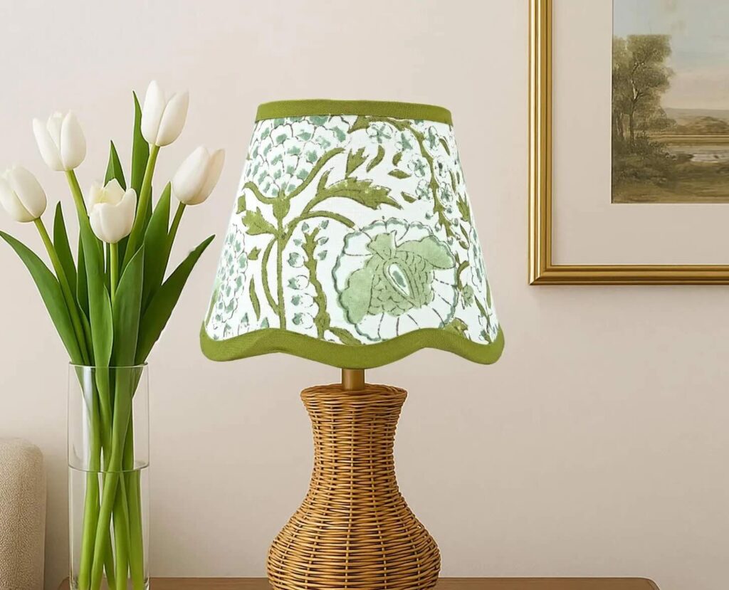 A wicker table lamp with a floral-patterned, green-trimmed lampshade sits next to a vase of white tulips; a framed landscape painting hangs on a light beige wall in the background.